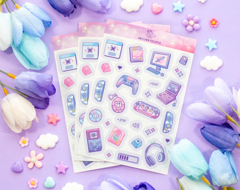 May include: A set of three clear sticker sheets with a pastel pink and purple background. The stickers feature a variety of cute cartoon illustrations of gaming consoles, controllers, and other gaming-related items. The text "Unicorn Eclipse" is printed on the top sheet.
