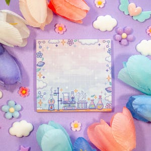May include: A square notepad with a science-themed border featuring beakers, flasks, and planets. The notepad has a grid pattern and a gradient background of pink, blue, and white. The image is surrounded by colorful flowers and decorative elements on a purple surface.