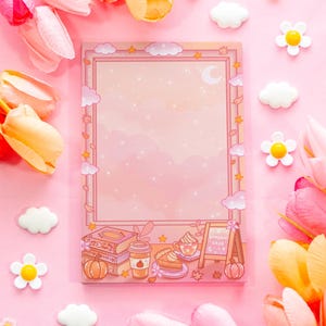 May include: A pink notepad with a decorative border featuring clouds, stars, and a crescent moon. The bottom of the notepad has illustrations of books, a pumpkin spice latte, pie, and a chalkboard. The notepad is surrounded by pink flowers and decorative accents.