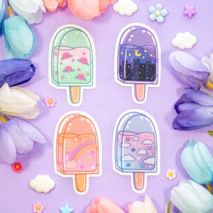 May include: Four colourful popsicle stickers with different designs. The stickers feature a green popsicle with pink flowers, a purple popsicle with a cityscape and moon, a pink popsicle with a rainbow and clouds, and a blue popsicle with clouds and stars.
