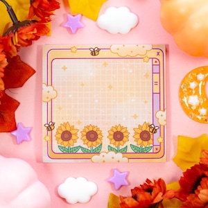May include: A yellow and white gridded notepad with a cute bee and sunflower design. The notepad has a purple border and is surrounded by fall leaves, a pumpkin, and other decorative elements.