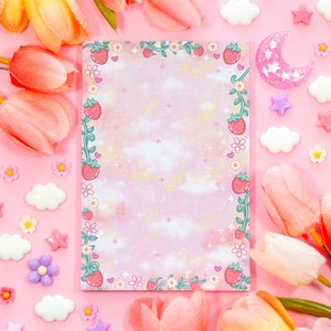 Könnte beinhalten: Ein rosa Notizblock mit Erdbeer- und Blumenrand, umgeben von dekorativen Elementen wie Tulpen, Wolken, Sternen und einem Mond. Der Notizblock hat einen weichen, wolkenartigen Hintergrund.