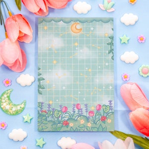 Könnte beinhalten: Ein blaues und grünes Notizbuch mit einem Gittermuster und einem Mond- und Sternen-Design. Das Notizbuch hat einen Rand aus Blumen und Blättern.