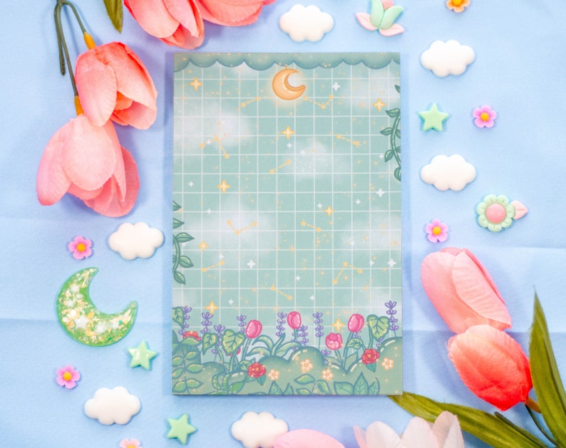 K&ouml;nnte beinhalten: Ein blaues und gr&uuml;nes Notizbuch mit einem Gittermuster und einem Mond- und Sternen-Design. Das Notizbuch hat einen Rand aus Blumen und Bl&auml;ttern.