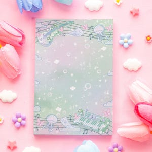 May include: A pastel-colored notepad with a musical theme. The design features a violin, piano, musical notes, and jellyfish illustrations. The notepad is surrounded by pink and white decorative elements, including flowers and clouds.