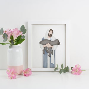 Puede incluir: Una ilustraci&oacute;n acuarela enmarcada de Jes&uacute;s sosteniendo un cordero. La ilustraci&oacute;n est&aacute; en un marco blanco y est&aacute; sobre una superficie blanca con flores rosas en un jarr&oacute;n blanco.
