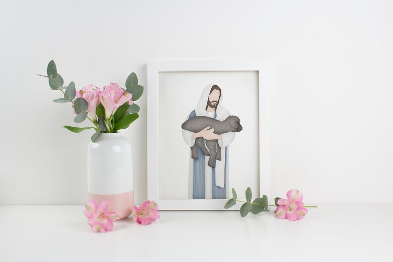 Puede incluir: Una ilustraci&oacute;n acuarela enmarcada de Jes&uacute;s sosteniendo un cordero. La ilustraci&oacute;n est&aacute; en un marco blanco y est&aacute; sobre una superficie blanca con flores rosas en un jarr&oacute;n blanco.