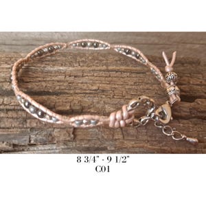 Könnte beinhalten: Ein hellrosa Wickelarmband mit dunkelgrauen Perlen und einem silbernen Verschluss. Das Armband misst 22.2 - 24.1 cm und wird auf einer Holzoberfläche präsentiert. Das Armband hat einen dekorativen Knoten und eine verstellbare Kette.