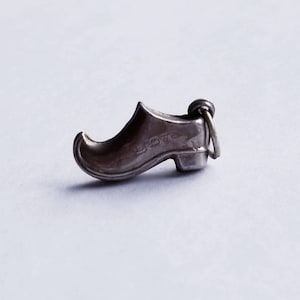 May include: A small, silver-colored charm in the shape of a wooden clog. The charm has a loop at the top for attaching to a bracelet or necklace. The word "LOVE" is engraved on the side of the clog. The charm is set against a light gray background.