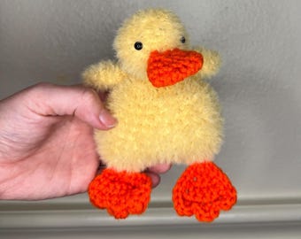 May include: A handmade yellow plush duck with orange feet and beak. The duck has small black eyes and a fluffy texture. The toy is designed to resemble a baby duckling.