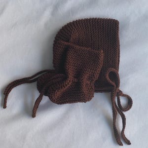 May include: A brown knitted baby bonnet and matching booties set. The bonnet has a rounded shape with a tie closure, and the booties are small and cozy. The set is made of a soft, textured yarn, perfect for a newborn.