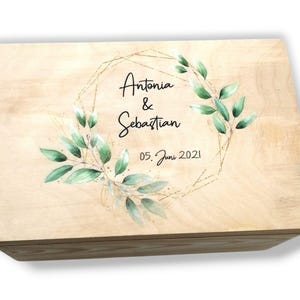 Könnte beinhalten: Eine hellbraune Holzkiste mit Deckel. Der Deckel zeigt die Namen "Antonia & Sebastian" und das Datum "05. Juni 2021", umrahmt von einem goldenen geometrischen Design und grünen Blättern.