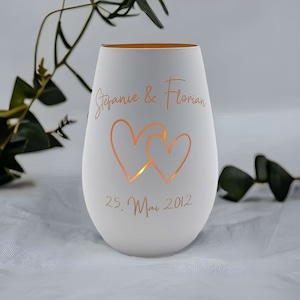 May include: White ceramic candle holder with two hearts cut out and the text "Stefanie & Florian 25. Mai 2012" in a gold script font.