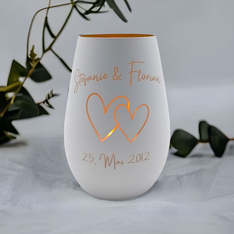May include: White ceramic candle holder with two hearts cut out and the text "Stefanie & Florian 25. Mai 2012" in a gold script font.