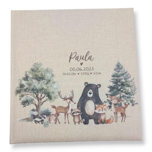 May include: A beige linen photo album with a woodland theme. The album features a brown bear, a deer, a raccoon, a fox, and a squirrel. The text "Paula" and "05.06.2023 14:42 Uhr 3210g 52cm" is printed on the front of the album.