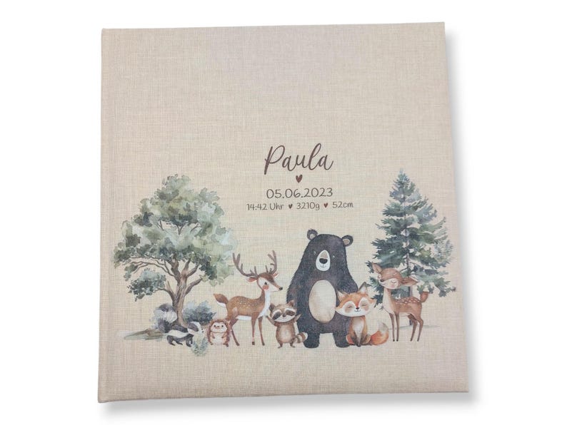 May include: A beige linen photo album with a woodland theme. The album features a brown bear, a deer, a raccoon, a fox, and a squirrel. The text "Paula" and "05.06.2023 14:42 Uhr 3210g 52cm" is printed on the front of the album.