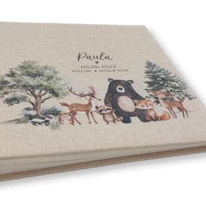 May include: A beige linen photo album with a woodland animal theme. The cover features a brown bear, a deer, a fox, a raccoon, and a skunk. The album is personalized with the name "Paula" and the date "05.06.2023".