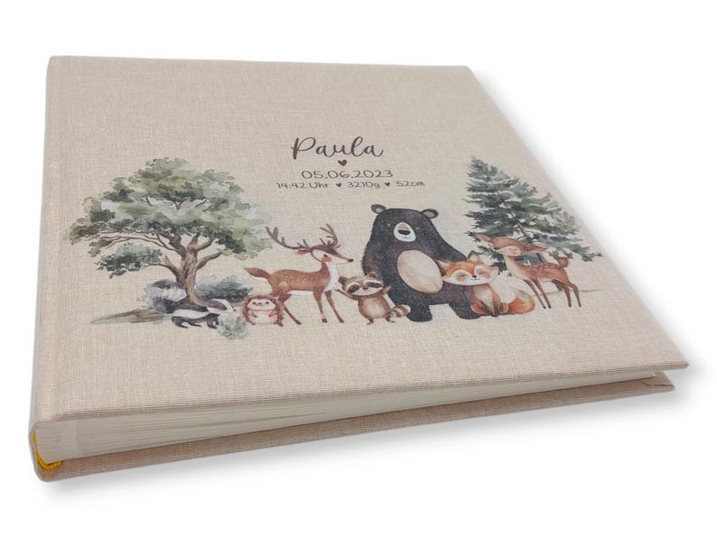 May include: A beige linen photo album with a woodland animal theme. The cover features a brown bear, a deer, a fox, a raccoon, and a skunk. The album is personalized with the name "Paula" and the date "05.06.2023".