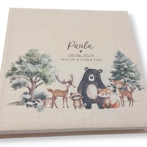 May include: A beige linen baby photo album with a woodland animal theme. The cover features a brown bear, a deer, a raccoon, a fox, a skunk, and a hedgehog. The album is personalized with the name "Paula" and the date "05.06.2023".