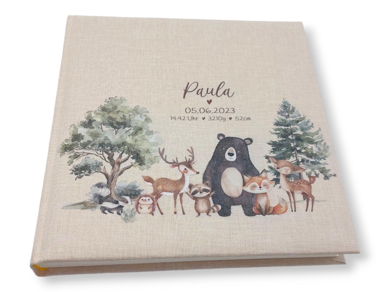 May include: A beige linen baby photo album with a woodland animal theme. The cover features a brown bear, a deer, a raccoon, a fox, a skunk, and a hedgehog. The album is personalized with the name "Paula" and the date "05.06.2023".