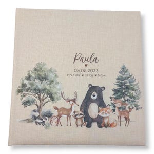 May include: A beige fabric baby photo album with a woodland animal theme. The album features a brown bear, a deer, a raccoon, a fox, and a squirrel. The text "Paula" is printed on the front of the album, along with the date "05.06.2023", the time "14:42 Uhr", the weight "3210g", and the length "52cm".