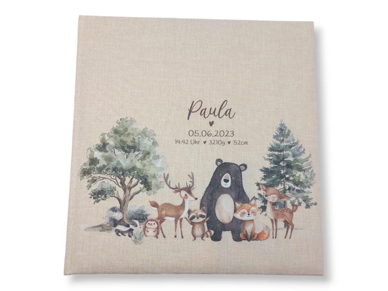 May include: A beige fabric baby photo album with a woodland animal theme. The album features a brown bear, a deer, a raccoon, a fox, and a squirrel. The text "Paula" is printed on the front of the album, along with the date "05.06.2023", the time "14:42 Uhr", the weight "3210g", and the length "52cm".