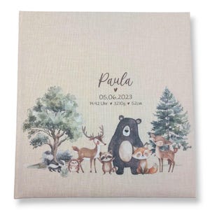 May include: A beige fabric photo album with a watercolor illustration of woodland animals, including a deer, a bear, a raccoon, a fox, and a bird. The album has the name "Paula" and the date "05.06.2023" printed on the front. The text "14:42 Uhr &hearts; 3210g &hearts; 52cm" is also printed on the front.