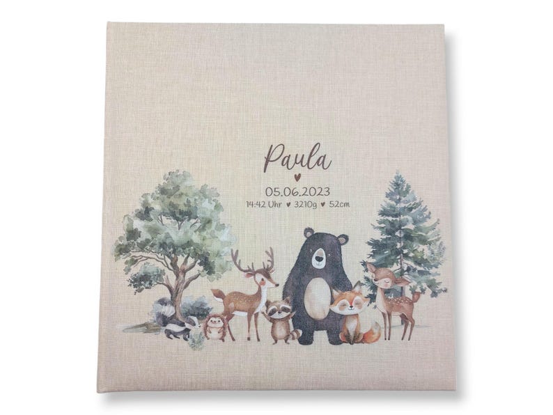 May include: A beige fabric photo album with a watercolor illustration of woodland animals, including a deer, a bear, a raccoon, a fox, and a bird. The album has the name "Paula" and the date "05.06.2023" printed on the front. The text "14:42 Uhr &hearts; 3210g &hearts; 52cm" is also printed on the front.