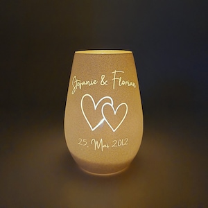 May include: A white glass candle holder with a frosted finish. The candle holder has two hearts and the text "Stefanie & Florian 25. Mai 2012" etched into the glass.