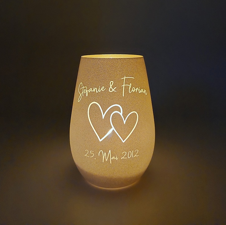 May include: A white glass candle holder with a frosted finish. The candle holder has two hearts and the text "Stefanie & Florian 25. Mai 2012" etched into the glass.