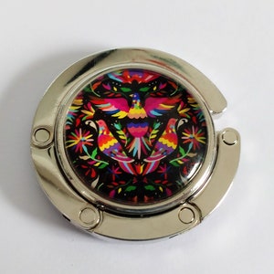 Peut inclure: Un crochet de sac en métal argenté avec un motif rond et coloré représentant des oiseaux et des fleurs. Le motif est sur fond noir.