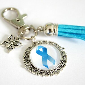 Può includere: Portachiavi argentato con un ciondolo a nastro blu, un ciondolo a farfalla e un nappa turchese. Il ciondolo rotondo presenta un disegno a nastro blu su sfondo bianco, incastonato in una cornice argentata ornata. Un accessorio elegante.