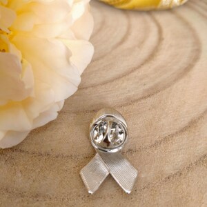 May include: Silver ribbon pin with a textured surface. The pin is in the shape of a ribbon with a loop at the top. The pin is on a wooden surface.