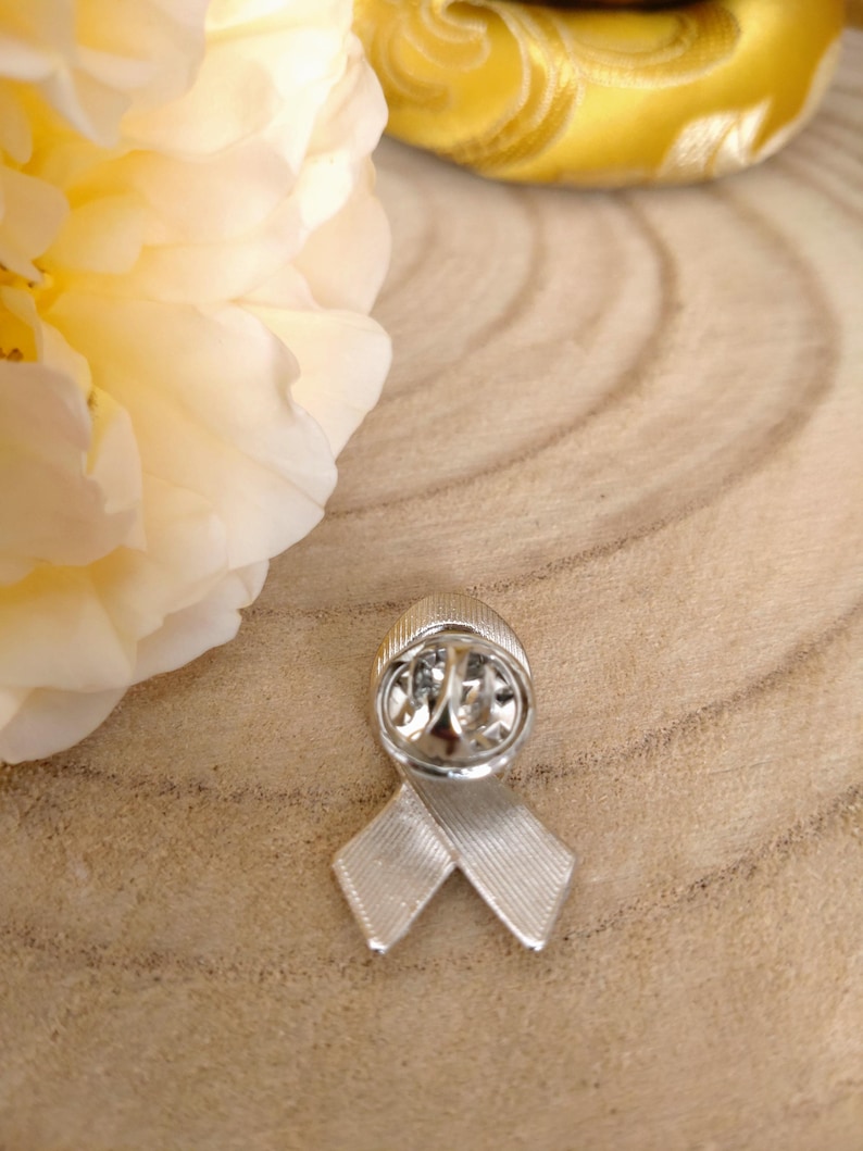 May include: Silver ribbon pin with a textured surface. The pin is in the shape of a ribbon with a loop at the top. The pin is on a wooden surface.