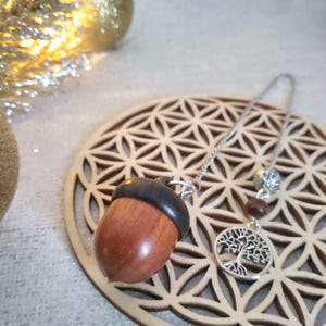 Op de afbeelding: Een houten eikelhanger met een donkerbruine dop en een zilveren ketting. De ketting leidt naar een zilveren levensboombedel en kleine kralen. De hanger ligt op een houten onderzetter met een geometrisch patroon.