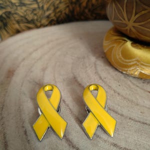 May include: Two yellow ribbon pins with a silver metal backing. The ribbons are tied in a bow shape.