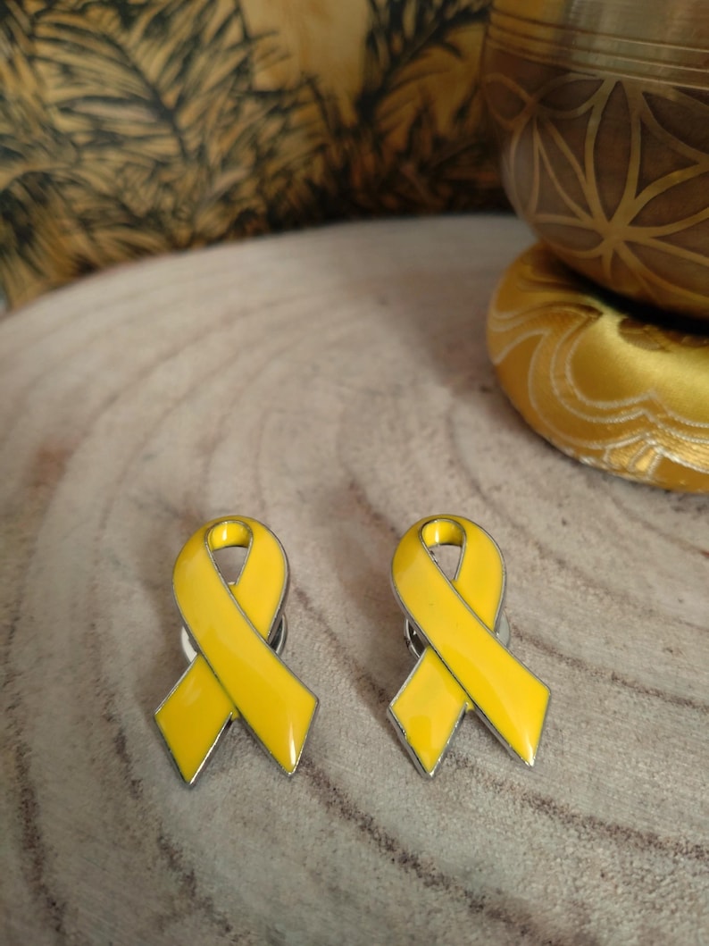 May include: Two yellow ribbon pins with a silver metal backing. The ribbons are tied in a bow shape.