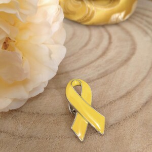 May include: A yellow enamel ribbon pin with a silver metal backing. The ribbon is tied in a bow shape.