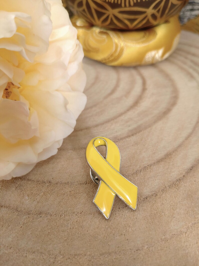 May include: A yellow enamel ribbon pin with a silver metal backing. The ribbon is tied in a bow shape.