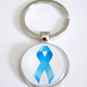 Può includere: Un portachiavi color argento con un ciondolo rotondo raffigurante un nastro azzurro chiaro su sfondo bianco. Il nastro è un simbolo di consapevolezza. Il portachiavi ha un anello argentato per attaccare le chiavi.