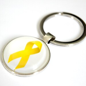 Può includere: Un portachiavi rotondo con un anello in metallo color argento e un ciondolo circolare. Il ciondolo presenta un nastro giallo su sfondo bianco, simbolo di consapevolezza. Un accessorio semplice ma significativo.