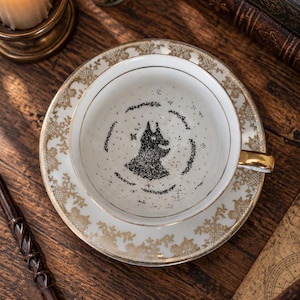 Pode incluir: Um conjunto de chávena e pires em porcelana branca com rebordo dourado e padrões intrincados de renda dourada. O interior da chávena apresenta uma silhueta preta pontilhada de uma criatura semelhante a uma raposa num motivo circular.