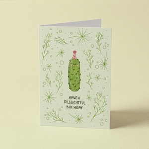 May include: A light green greeting card featuring a central illustration of a smiling pickle wearing a pink party hat. The background is decorated with green dill sprigs and small circles. The text reads: HAVE A DILL-LIGHTFUL BIRTHDAY.