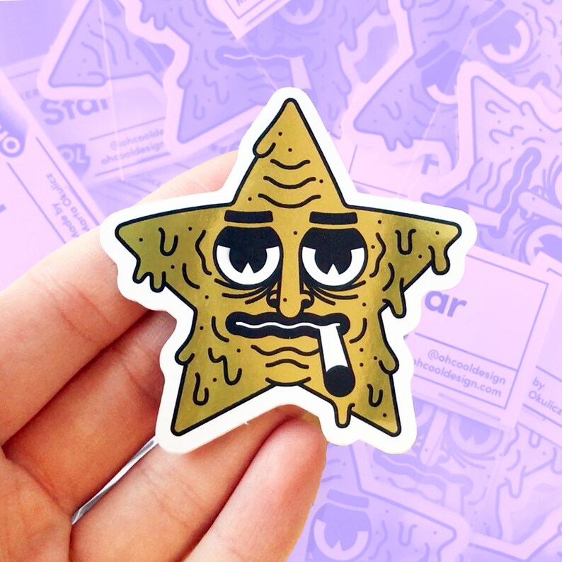 Op de afbeelding: Een gouden folie sticker van een cartoonster met een menselijk gezicht, met een serieuze uitdrukking en een sigaret roken. De ster druipt met gouden vloeistof. De sticker zit op een hand.
