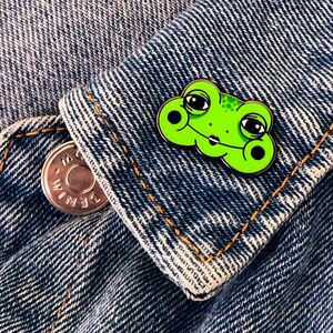 May include: Green enamel pin of a frog with large eyes and a closed mouth. The frog is wearing a light pink lipstick. The pin is on a blue denim jacket.