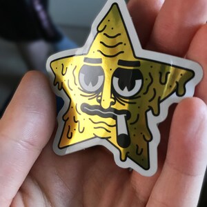 Op de afbeelding: Een gouden folie sticker van een cartoonster met een smeltend gezicht en een sigaret in zijn mond.