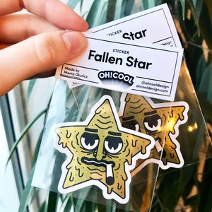 Op de afbeelding: Twee gele stickers in de vorm van een ster met een cartoon gezicht, hangende ogen en een sigaret in de mond. De stickers zijn verpakt in een doorzichtige plastic zak met een wit label waarop "Fallen Star" en "OH!COOL" staat.