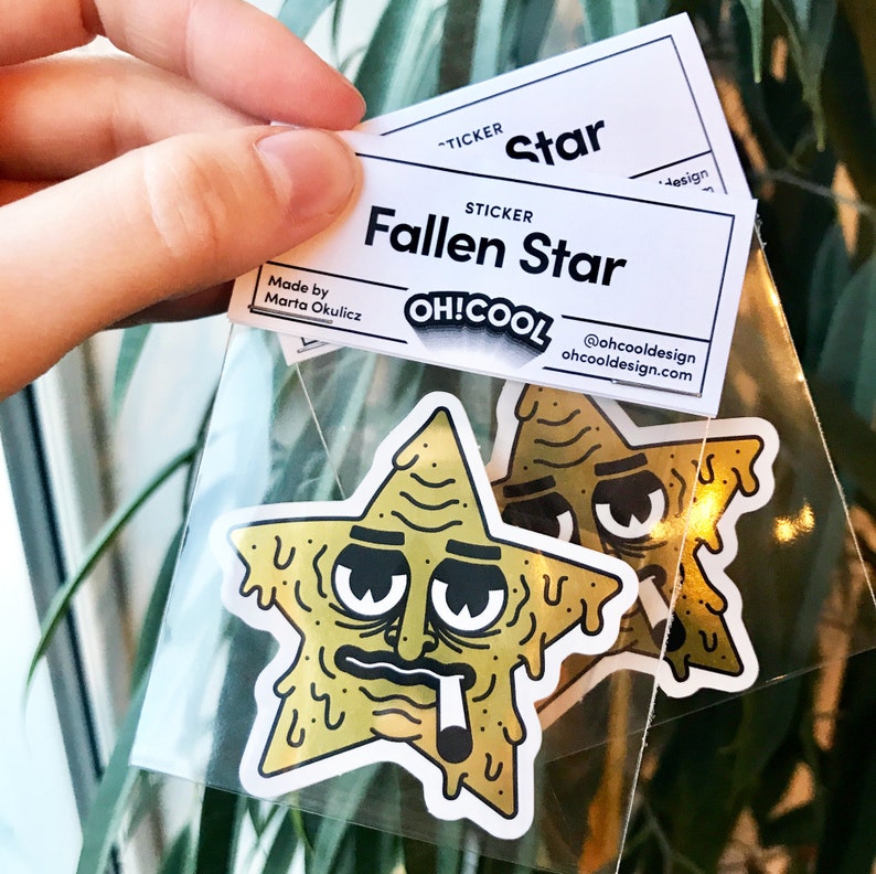 Op de afbeelding: Twee gele stickers in de vorm van een ster met een cartoon gezicht, hangende ogen en een sigaret in de mond. De stickers zijn verpakt in een doorzichtige plastic zak met een wit label waarop "Fallen Star" en "OH!COOL" staat.