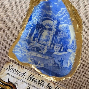 May include: A blue and white porcelain dish with a gold rim. The dish features a vintage design of a building with a bridge and trees. The dish is sitting on a burlap surface. The text "Sacred Hearts by Kim" is visible on a card in the foreground. Mixed media art and vintage treasures.