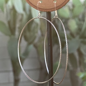 May include: A pair of silver hoop earrings. One earring has a smooth, polished surface, while the other features a textured, dotted design. The earrings are suspended from delicate ear wires and displayed on a wooden stand against a blurred green background.